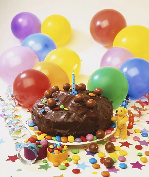 Small chocolate cake decorated with sweets and a lit candle, surrounded by colourful balloons, scattered sweets, paper stars and ribbons