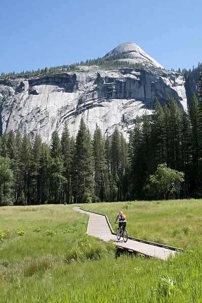 USA, California, Yosemite Valley, woman cycling on boardwalk through lush countryside with mountain in background