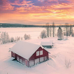 Europe, Finland, Rovaniemi, aerial view of a frozen lake near Rovaniemi with traditional red buildings with the sun rising on a winter morning