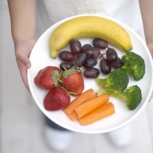 Girl holding plate of fruit and vegetables, including banana, strawberries, grapes, broccoli, carrots