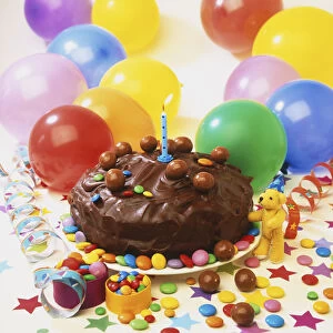 Small chocolate cake decorated with sweets and a lit candle, surrounded by colourful balloons, scattered sweets, paper stars and ribbons