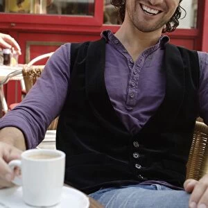 Man, early 30s, wearing casual clothes sitting in an outdoor cafAzA with a coffee cup, smiling